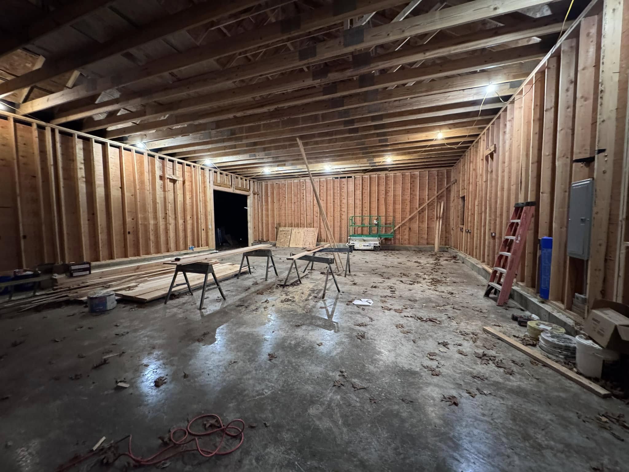 Large Space ready to be remodeled in Columbia Connecticut Large unfinished interior space with exposed wooden framing and open ceiling joists, prepared for remodeling. The area features a concrete floor, scattered construction tools, sawhorses, a red ladder, and stacked lumber. Overhead work lights illuminate the structure. Located in Columbia, Connecticut.